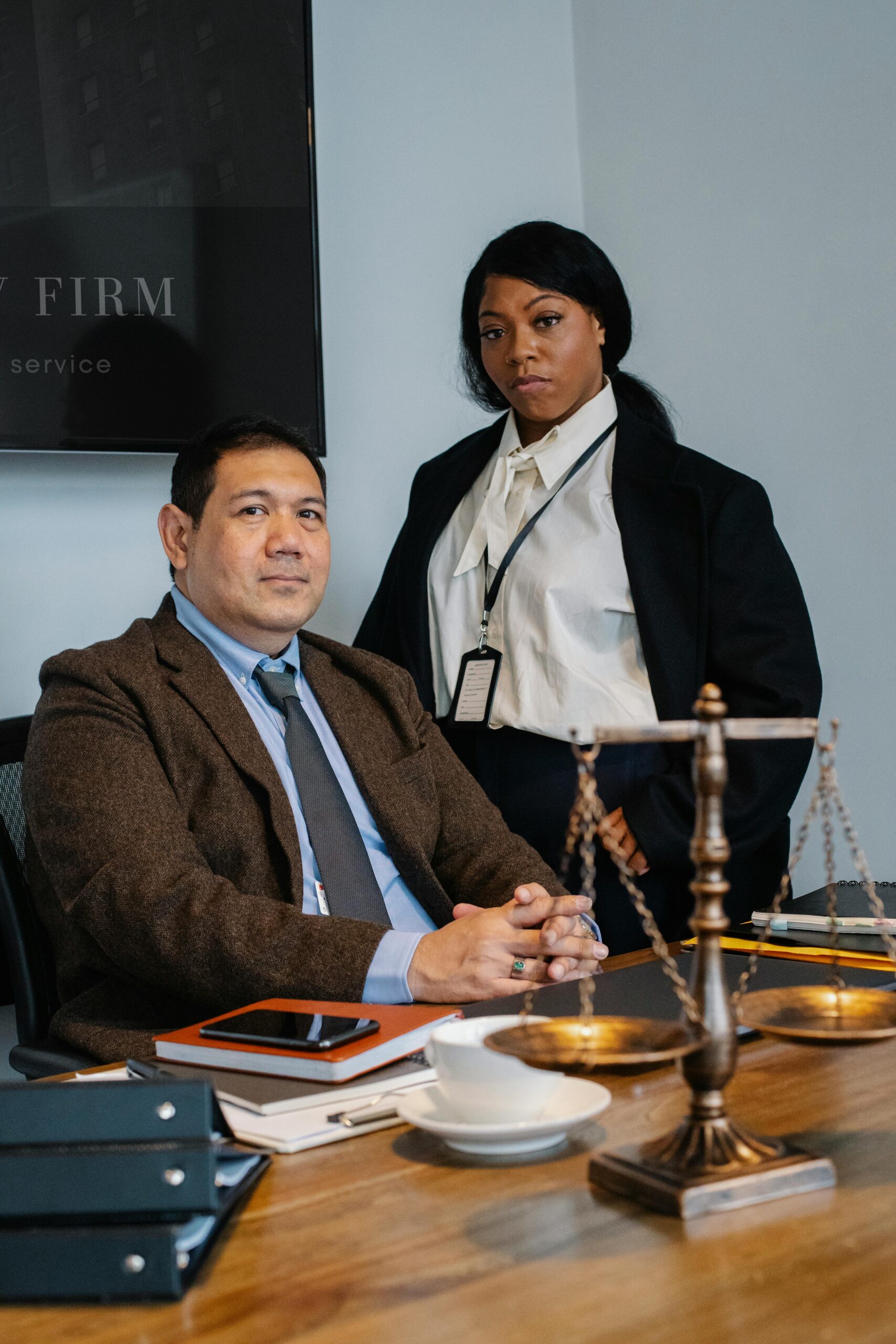 Focused young black woman in formal clothes standing near male ethnic colleague at wooden table with documents and justice scale during work in law firm